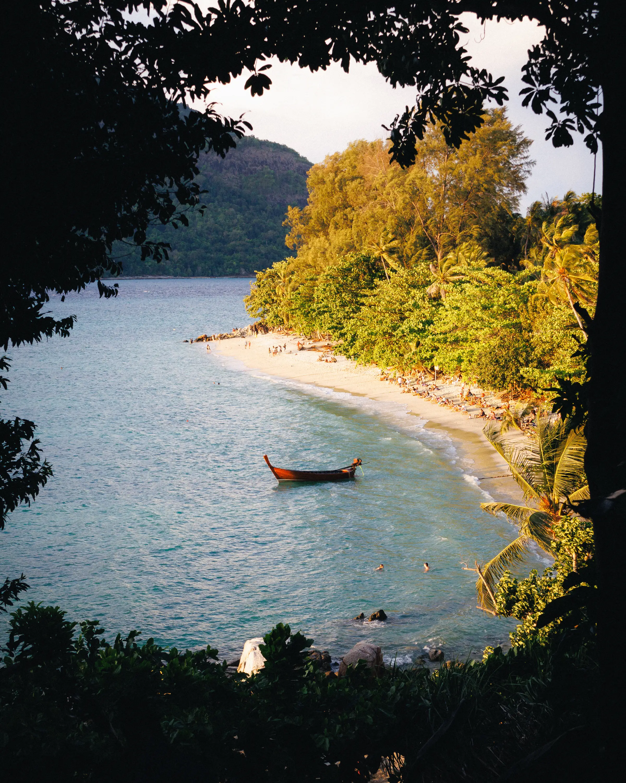 A longtail boat anchored in turquoise water off a curved beach, seen through dark leaves